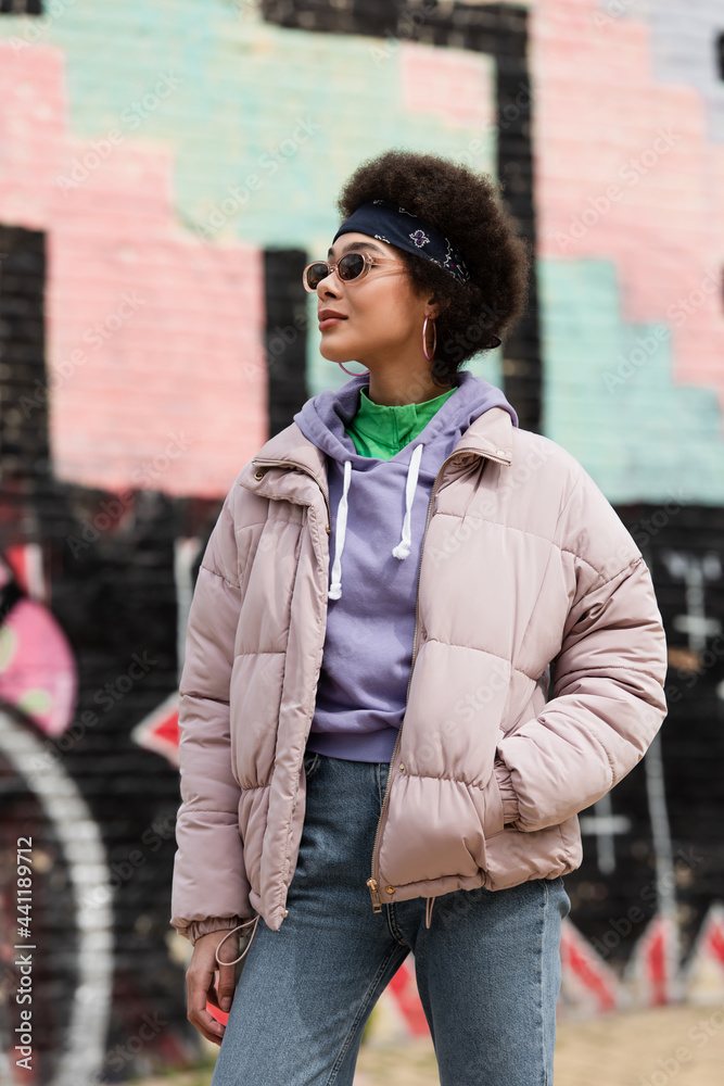 African american woman in sunglasses holding hand in pocket of jacket on urban street