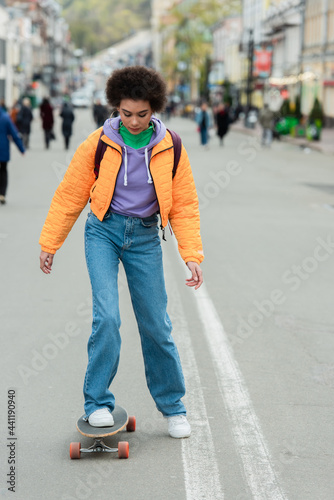 Young african american woman with backpack riding longboard on urban street