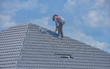 A construction worker wearing a full body safety belt is installing tile on the roof of a large house. By using corrugated ceramic tiles on a strong steel roof frame.