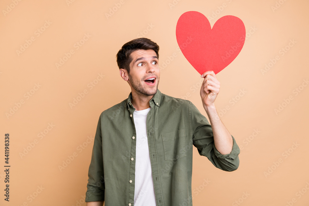 Photo of amazed charming happy man look empty space hold red paper heart isolated on beige color background Photo of amazed charming happy man look empty space hold red paper heart isolated on beige color background