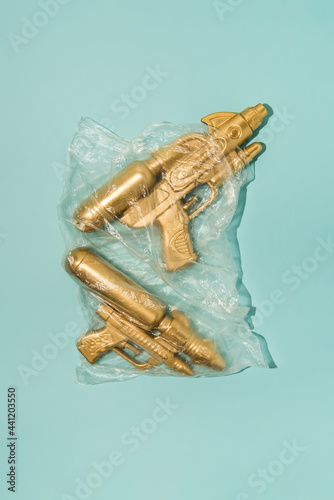 Golden water-gun wrapped in transparent plastic bag on pastel blue background. Creative summer concept with copy space.