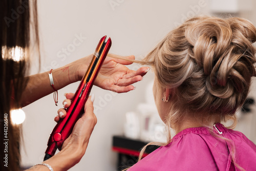 A girl-hairdresser makes a client's hair for a celebration with the help of an iron-straightener.