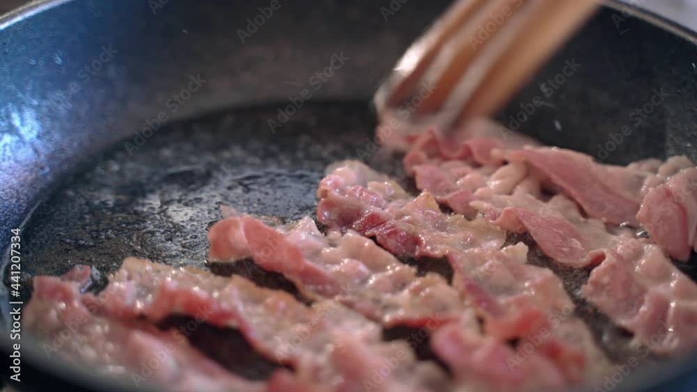 Roasted fresh bacon strips on hot pan wih sizzling oil, process of meat slices frying at home for tasty morning breakfast. Person using wooden fork for turning around bacon strips while frilling on