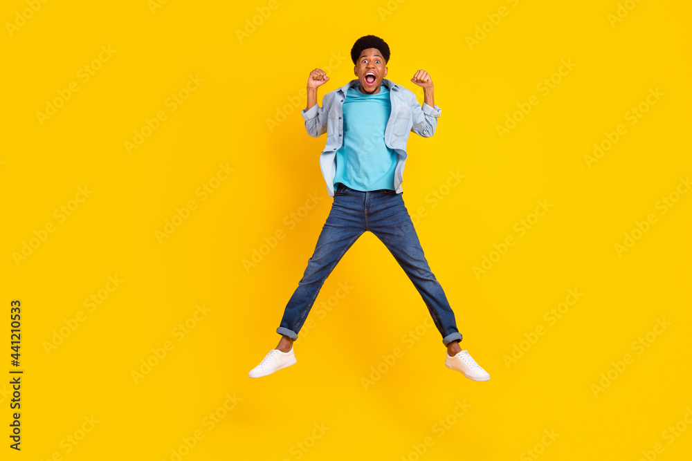 Photo of sweet lucky dark skin man wear jeans shirt jumping high rising fists smiling isolated yellow color background