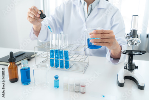 Scientist woman holding beaker and dropper to dropping blue liquid into test tube while working with microscope