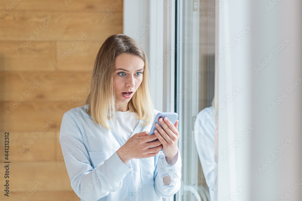 young woman saw bad news on smartphone, standing by the window in the office or at home indoors. The female anxiously holds a mobile phone in her hands, reads the news of the day young woman saw bad news on smartphone, standing by the window in the office or at home indoors. The female anxiously holds a mobile phone in her hands, reads the news of the day