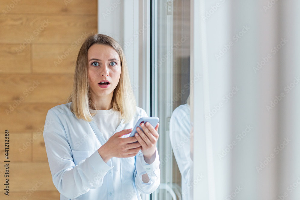 young woman saw bad news on smartphone, standing by the window in the office or at home indoors. The female anxiously holds a mobile phone in her hands, reads the news of the day young woman saw bad news on smartphone, standing by the window in the office or at home indoors. The female anxiously holds a mobile phone in her hands, reads the news of the day