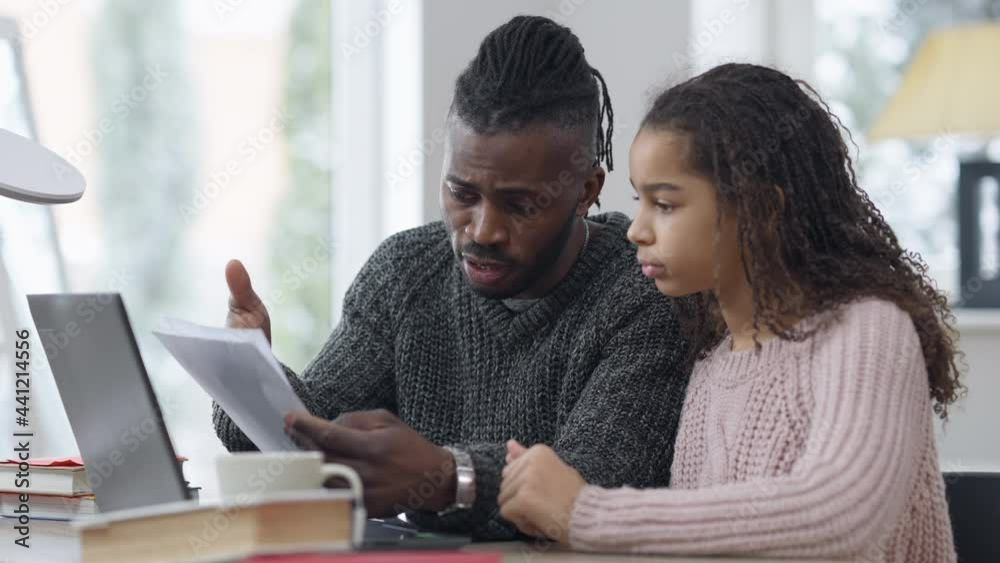 Handsome African American father helping cute adolescent daughter with homework. Portrait of intelligent serious man talking explaining to pretty schoolgirl sitting at table indoors. Family support Handsome African American father helping cute adolescent daughter with homework. Portrait of intelligent serious man talking explaining to pretty schoolgirl sitting at table indoors. Family support