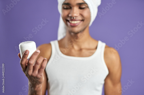 Young man smiling with cosmetic face cream in hand on purple background close up portrait