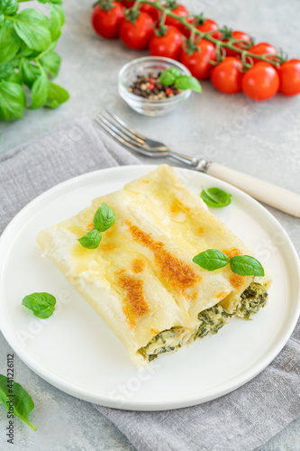 Traditional Italian cannelloni with ricotta and spinach in bechamel sauce with fresh basil leaves on a white plate on a gray concrete background. Vegetarian dish. Copy space.