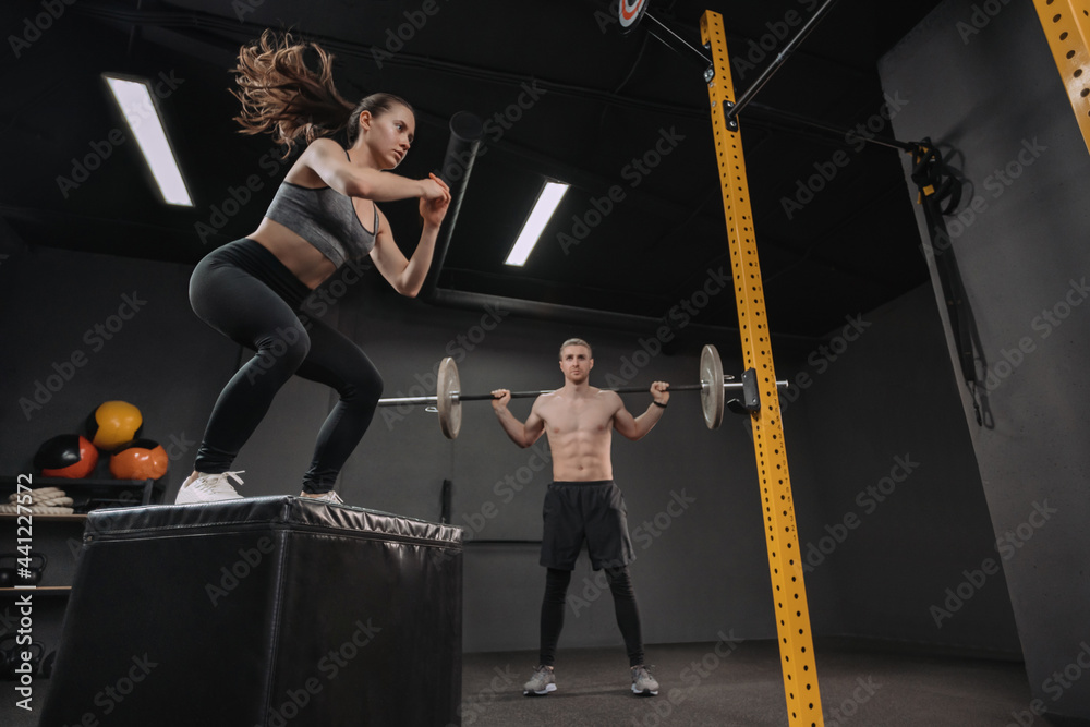 Two young crossfit athletes exercising together at gym. Sporty fit couple having interval functional training class. Pretty female doing box jump exercise while muscular male doing back squats with ba