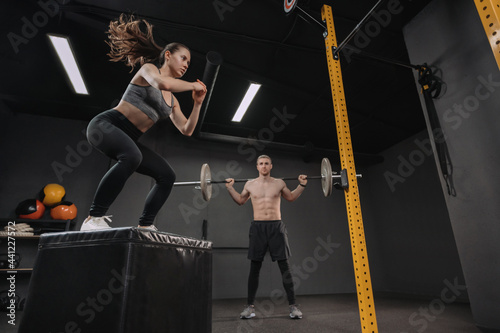 Two young crossfit athletes exercising together at gym. Sporty fit couple having interval functional training class. Pretty female doing box jump exercise while muscular male doing back squats with ba