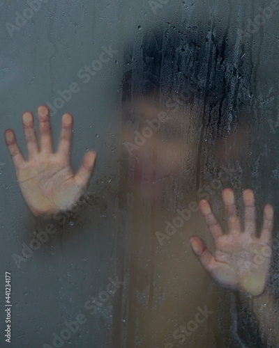 silhouette of child washing suffering behind wet glass in shower crying with hands resting on the glass partition