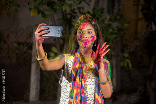 Young girl with colorful face taking selfie using smartphone on Holi festival. Festival and technology concept