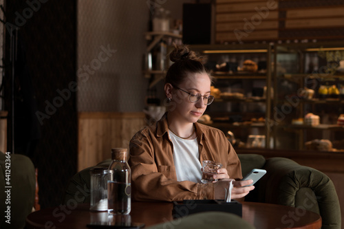 pretty young woman drinking water in cafe and wait for ordered food, using mobile phone for chatting or texting or surfing the internet
