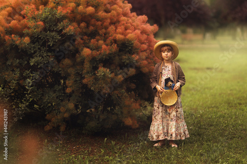 a girl in a hat with a toy teddy bear in her hands is standing in a beautiful summer garden