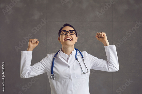Fotografía Young female doctor sincerely rejoices raising fists and smiling on a gray background