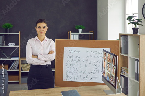 Portrait of confident serious young female teacher with folded arms on chest standing near board and desk with opened video conference chat on computer screen looking at camera. Online education