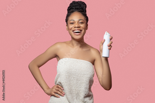 Dark-skinned young woman spraying deodorant on her body after shower