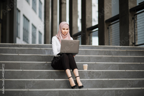 Charming woman in stylish elegant clothes and hijab sitting on stairs outdoors and using wireless laptop. Muslim female working on distance. © sofiko14