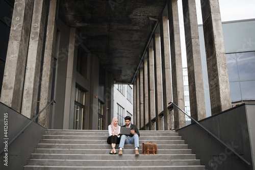 Handsome man and beautiful woman working on wireless laptop near office center. Two arabian colleagues sitting on stairs and doing remote work.