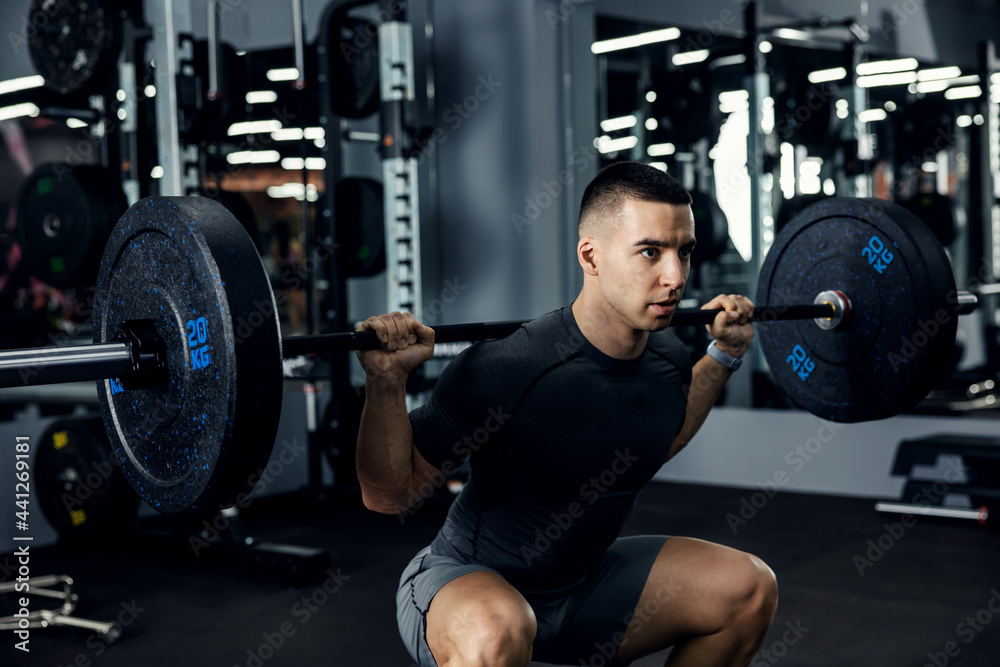 Close up side portrait of a male fitness trainer in grey sportswear doing squats using a barbell in a gym to train his legs and back. Leg burning workout, sports lover Close up side portrait of a male fitness trainer in grey sportswear doing squats using a barbell in a gym to train his legs and back. Leg burning workout, sports lover