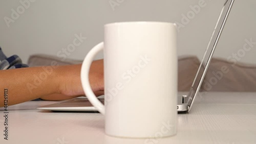A young woman drinks coffee from a white cup and types on a laptop keyboard. The concept of freelancing and working from home.