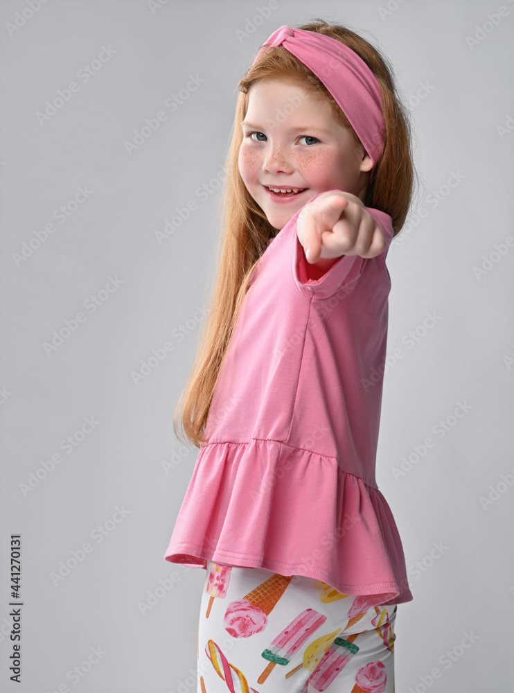 Friendly widely smiling little red-haired girl child wearing summer fashion outfit turning from back pointing to camera. Charming small female model studio portrait shot. Positive children emotion Friendly widely smiling little red-haired girl child wearing summer fashion outfit turning from back pointing to camera. Charming small female model studio portrait shot. Positive children emotion