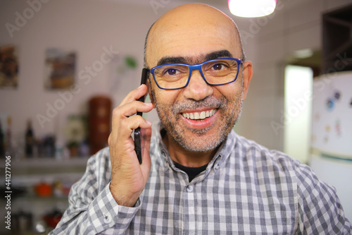 Homem de 50 anos dentro de casa olhando para a tela do celular conversando. photo