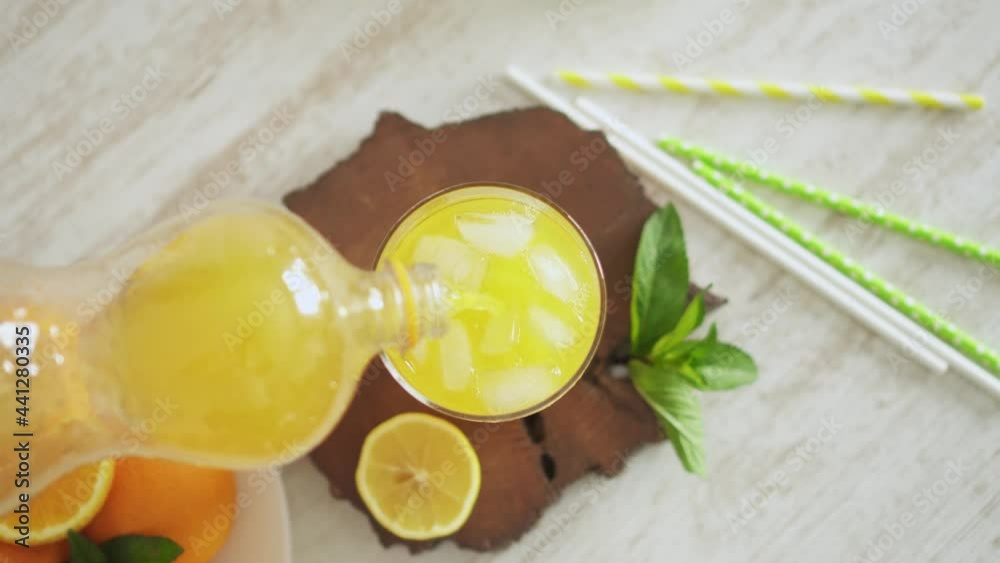 Orange soft drink from a plastic bottle is poured into a glass with ice cubes. A glass with a refreshing summer drink on a white kitchen table. Oranges, lemons and cocktail rolls. View from above.
