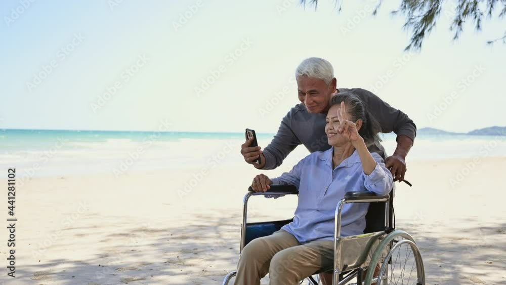 Asian senior couple tourist smile and pose by hands symbol to camera video call and make selfie by smartphone at beach. Couple elderly retire rest and relax wife is disabled and sitting on wheelchair. Asian senior couple tourist smile and pose by hands symbol to camera video call and make selfie by smartphone at beach. Couple elderly retire rest and relax wife is disabled and sitting on wheelchair.