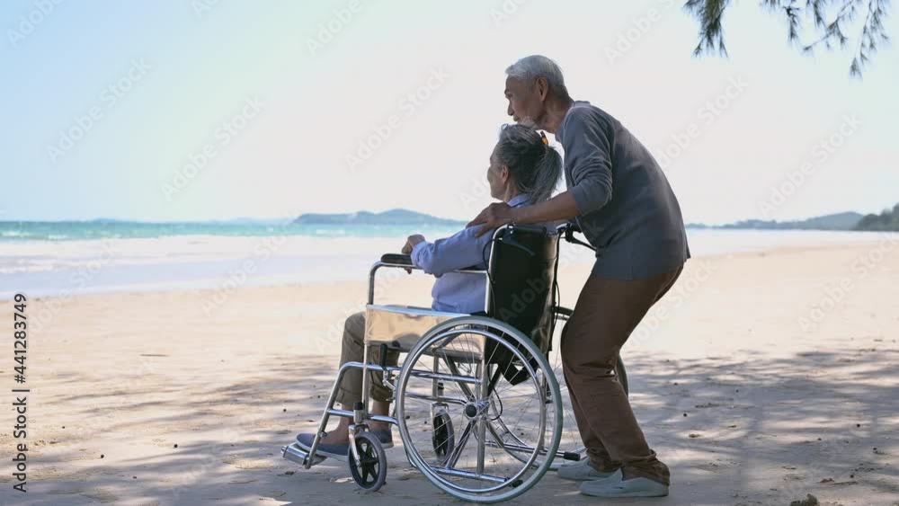 Side view of asian senior couple relaxing smile and enjoying with pointing and look to sea on beach under tree. Happy couple elderly retire resting and relax wife is disabled and sitting on wheelchair Side view of asian senior couple relaxing smile and enjoying with pointing and look to sea on beach under tree. Happy couple elderly retire resting and relax wife is disabled and sitting on wheelchair