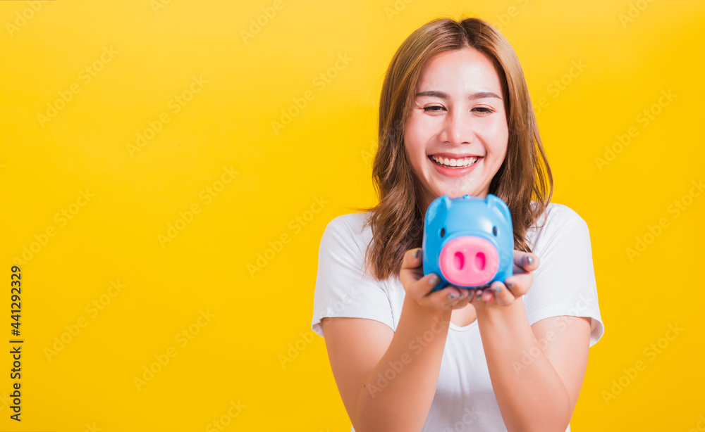 Portrait Asian Thai beautiful happy young woman smiling holding blue piggy bank with a lot of money and looking to piggybank, studio shot isolated on yellow background, with copy space Portrait Asian Thai beautiful happy young woman smiling holding blue piggy bank with a lot of money and looking to piggybank, studio shot isolated on yellow background, with copy space