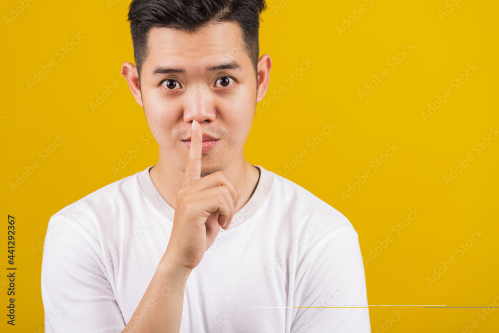 Asian handsome young man holding index finger on lips asking for silence or secret gesturing, studio shot isolated on yellow background, making shh shush gesture concept Asian handsome young man holding index finger on lips asking for silence or secret gesturing, studio shot isolated on yellow background, making shh shush gesture concept