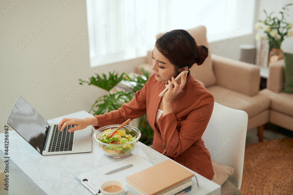 Pretty young woman eating healthy salad in office while talking on mobile phone