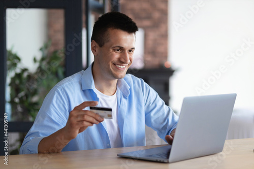 Happy smiling young male or student with a credit card using laptop for profitable purchases in the Internet sitting at home, online shopping