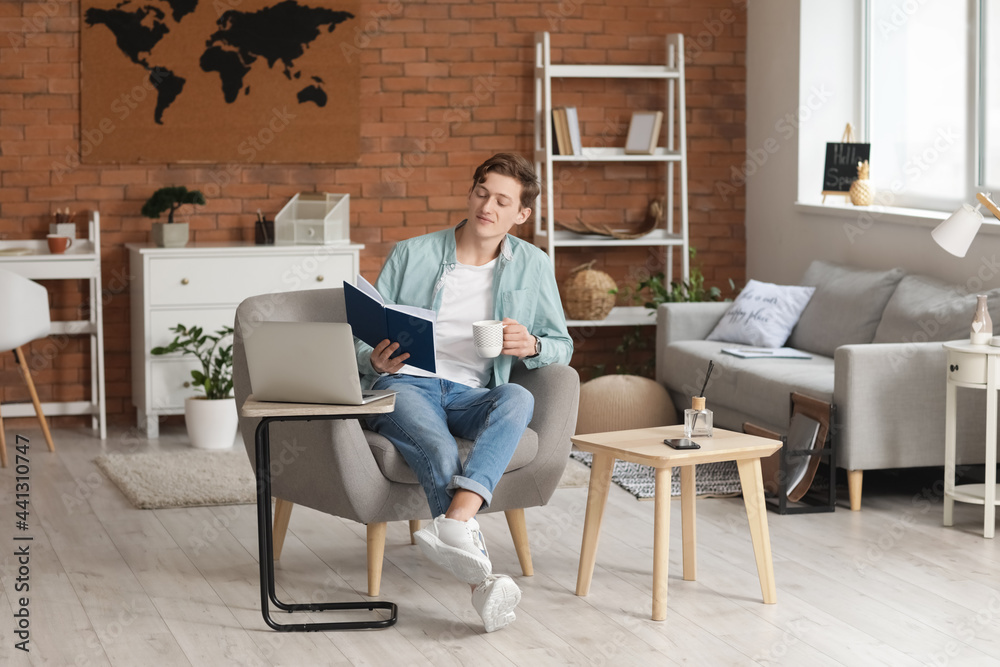 Young man reading book and drinking coffee while sitting in armchair at home