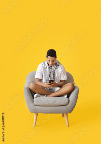 Young African-American guy with phone sitting in armchair on color background © Pixel-Shot