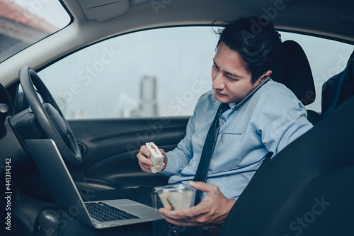 Businessman eating sandwich while working in laptop and talking on the phone  in the drivers seat in his car. Busy businessman and food in car.