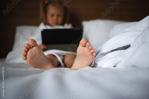 Little cute girl in a white robe looks at the tablet while lying on the bed