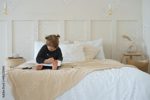 a little girl in black home clothes is sitting on the bed and drawing with felt-tip pens
