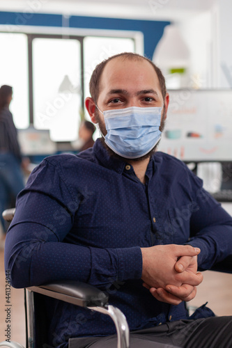 Employee with disabilities, invalid, handicapped paralized with face mask against coronavirus looking at camera sitting immobilized in wheelchair in business office room, working at financial project. photo