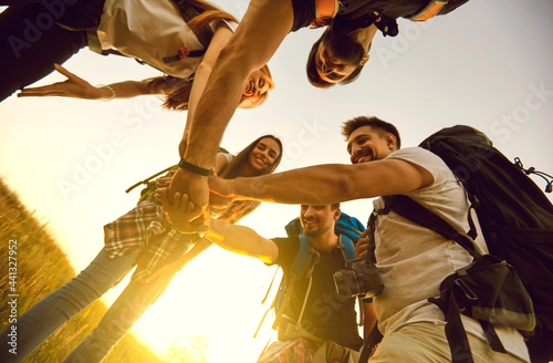 From below of a team of happy young friends joining hands ready for great travel adventure. Low angle of group of tourists with backpacks stacking hands in summer field while hiking in countryside