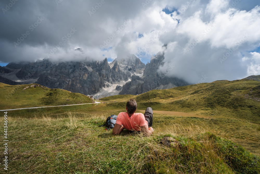 Dolomites. Man laying on the grass enjoying Baita Segantini mountain with Cimon della Pala peak, refuge and lake in background. Rolle pass, Trentino province, Italy, Europe