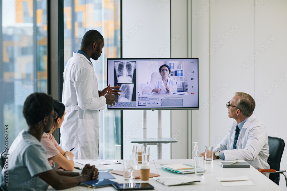 Obraz premium Portrait of African-American doctor standing by digital screen in onference room and talking to expert via video call, copy space