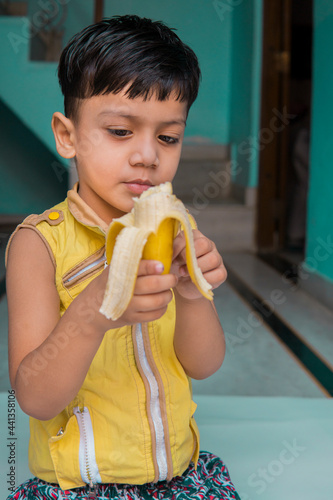 Indoor image of an Asian, Indian cute little boy in yellow clothes eating a fresh banana at home. 