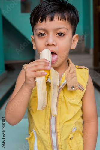 Indoor image of an Asian  Indian cute little boy in yellow clothes eating a fresh banana at home. 