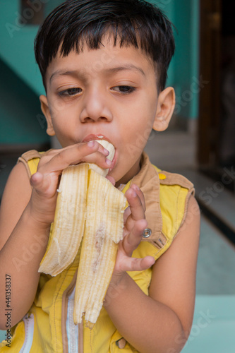 Indoor image of an Asian  Indian cute little boy in yellow clothes eating a fresh banana at home. 