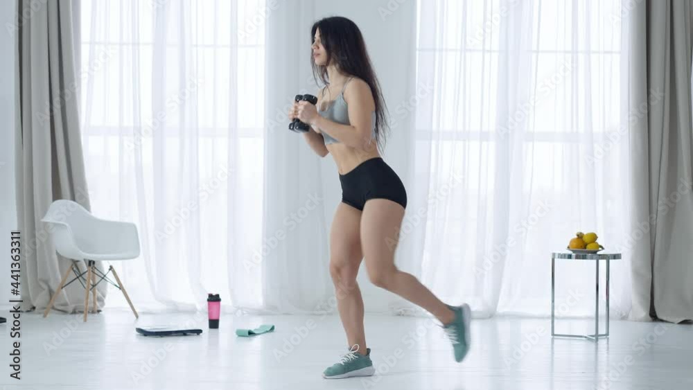 Side view of slim woman doing single-leg squats with dumbbells in hands. Wide shot of fit athletic sportive Middle Eastern sportswoman training indoors at home. Athleticism and sport concept Side view of slim woman doing single-leg squats with dumbbells in hands. Wide shot of fit athletic sportive Middle Eastern sportswoman training indoors at home. Athleticism and sport concept