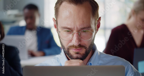 Close up of young businessman sitting at desk working on laptop and looking thoughtfully in office.